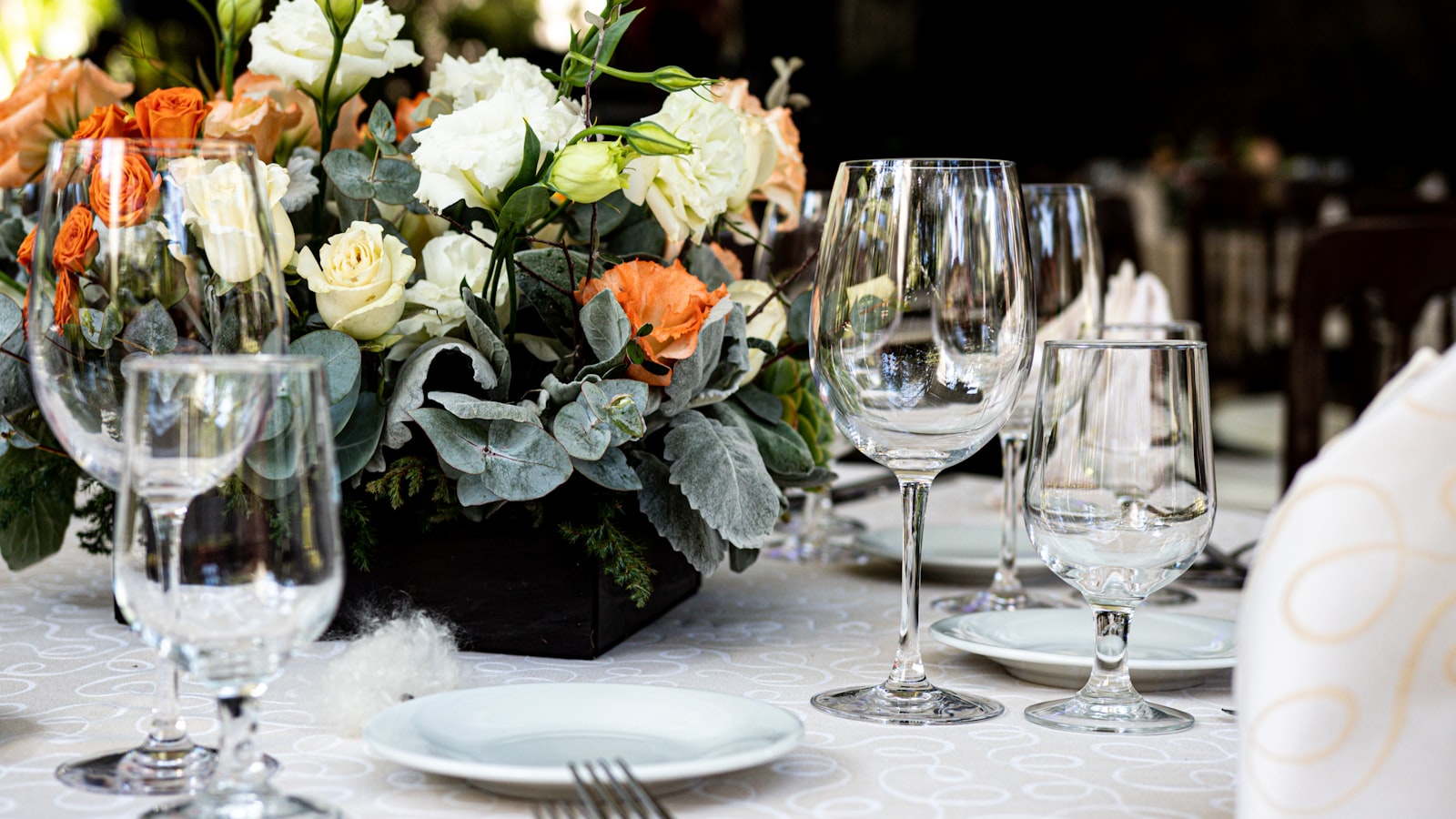 Round wedding reception table set with white linen, gold flatware, and white and orange floral centerpieces